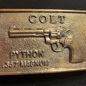 May include: A rectangular brass-toned metal belt buckle featuring a raised relief of a Colt Python revolver. The text COLT is embossed at the top, with PYTHON and .357 MAGNUM embossed at the bottom. The buckle is attached to a dark brown leather belt.