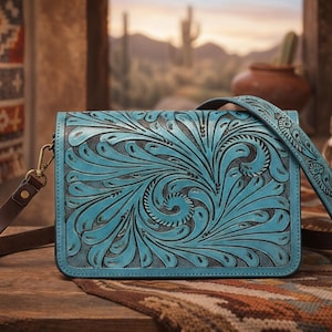 May include: A turquoise leather crossbody bag with a detailed floral pattern. The bag has a brown leather strap and gold-colored hardware. The background includes a Southwestern-style rug and a desert landscape.