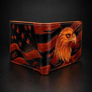 May include: A bi-fold leather wallet featuring a hand-tooled design of a bald eagle head in shades of brown and gold, set against a background of a stylized American flag with black and brown stripes and carved stars on the left side.