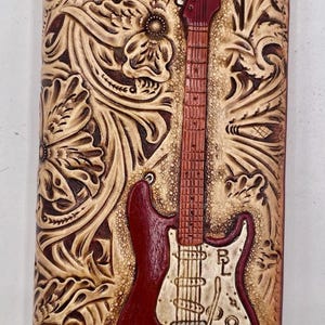 May include: A leather wallet featuring a carved floral pattern and a red electric guitar. The guitar is centrally placed, with the initials "RL" visible. The wallet is a deep brown colour.