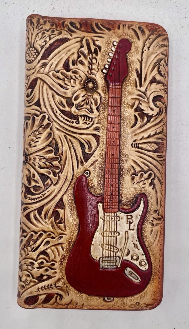 May include: A leather wallet featuring a carved floral pattern and a red electric guitar. The guitar is centrally placed, with the initials "RL" visible. The wallet is a deep brown colour.