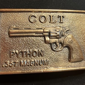 May include: A rectangular, bronze-colored belt buckle featuring a detailed relief of a Colt Python .357 Magnum revolver. The word "COLT" is at the top, with "PYTHON" and ".357 MAGNUM" below the gun.