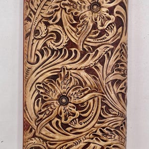 May include: A brown leather wallet with a floral design. The wallet features a detailed embossed pattern of flowers and leaves. The design is in a lighter tan colour, contrasting with the darker brown background. The wallet is rectangular and appears to be closed.