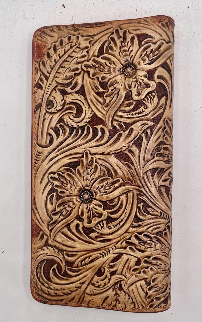 May include: A brown leather wallet with a floral design. The wallet features a detailed embossed pattern of flowers and leaves. The design is in a lighter tan colour, contrasting with the darker brown background. The wallet is rectangular and appears to be closed.