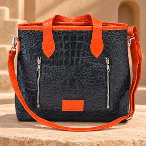 May include: A black tote bag with a textured crocodile skin pattern, featuring bright orange leather handles, trim, and a rectangular patch. The bag includes two vertical silver zippered pockets on the front and a detachable orange shoulder strap.