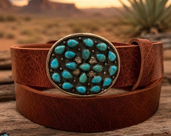 May include: A brown leather belt featuring an oval-shaped metal buckle. The buckle is decorated with numerous small, teardrop-shaped turquoise stones and small metal leaf accents arranged in a cluster across the dark, textured metal surface.