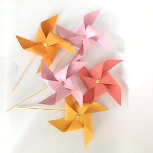 May include: Five colorful paper pinwheels with yellow centers and wooden sticks. The pinwheels are orange, pink, and red.