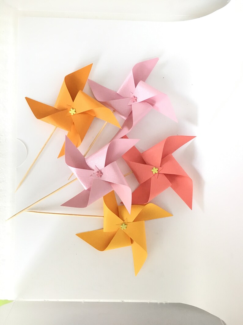May include: Five colorful paper pinwheels with yellow centers and wooden sticks. The pinwheels are orange, pink, and red.