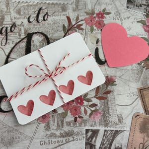 May include: A white card with three red watercolor hearts tied with red and white twine. A pink heart-shaped cutout and floral and map-themed paper are in the background. The word "to" is visible in the background.