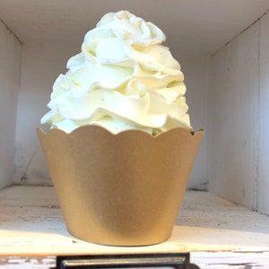 May include: A cupcake with a generous swirl of white frosting sits in a brown paper cup with a scalloped edge. The cupcake is displayed in a white, rustic-style shelf.
