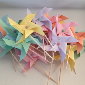 May include: A group of colorful paper pinwheels with pastel colors, including pink, blue, yellow, and green. Each pinwheel has a small, round, colorful button in the center. The pinwheels are attached to wooden skewers.