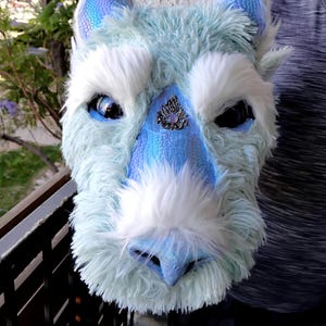 Op de afbeelding: Een fantasie wezenmasker in blauwe en witte tinten. Het masker heeft een getextureerde snuit en oren, met pluizig wit haar rond de ogen en snuit. Een decoratief juweel is op het voorhoofd bevestigd.