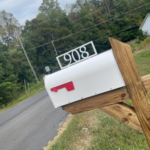 Puede incluir: Un buzón blanco con una bandera roja y el número 908 en un letrero rectangular blanco. El buzón está montado en un poste de madera, con una carretera y árboles al fondo.