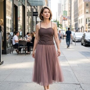 May include: A brown sleeveless top with a subtle white polka dot pattern and thin straps paired with a mauve-colored midi-length tulle skirt. The skirt features a layered, sheer fabric design that falls below the knees, complemented by light pink shoes.