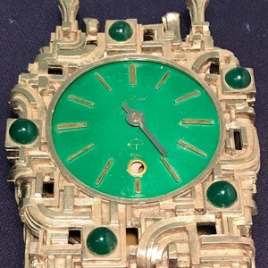 Può includere: Un orologio dorato con un quadrante verde e numeri romani. L'orologio ha un design unico e astratto con più livelli e accenti di pietre preziose verdi. L'orologio è a 8 giorni e realizzato in Germania.