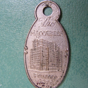 Peut inclure: Un porte-clés en argent avec un motif de bâtiment et le texte "The Maccabees Founded 1878".