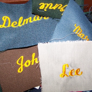 May include: Close-up of fabric patches with names embroidered in yellow cursive script. The patches are various colours, including blue, brown, green, and white. The names include "Delmar", "John", "Lee", and "Max".