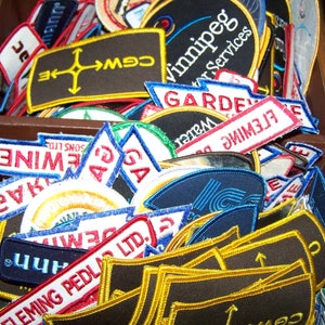 Op de afbeelding: Een close-up van een stapel kleurrijke stoffen patches met verschillende tekst en ontwerpen. De patches zijn gemaakt van verschillende materialen en hebben een verscheidenheid aan kleuren, waaronder rood, blauw, geel, groen en zwart. Sommige van de patches hebben tekst erop, zoals "Winnipeg Car Services", "Fleming Pedla Ltd" en "CGW".