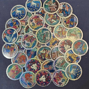 May include: A collection of iridescent, circular tokens featuring various whimsical designs. The tokens showcase images of unicorns, teddy bears, and other fantasy creatures, with vibrant colors and intricate details. The tokens are arranged on a dark blue surface.
