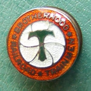 May include: A round, orange and white enamel pin with the text "Brotherhood of Railroad Trainmen" and a green "T" in the center.