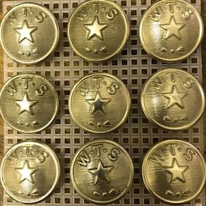 Puede incluir: Nueve botones de metal de color dorado con las letras "W.T.S." y una estrella de cinco puntas en el centro.