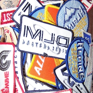 May include: A collection of colourful embroidered patches with various designs and text, including "MLJ", "ALL", "Alpine Messenger Service LTD", and "First", on a white background.