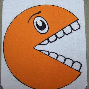 以下が含まれることがあります： 白いキャンバスに描かれた、口を大きく開けて白い歯を見せたオレンジ色のパックマンのキャラクターです。