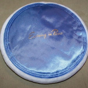May include: A round, blue satin powder puff with a white, fluffy edge. The puff has the words "Evening in Paris" written in gold lettering.