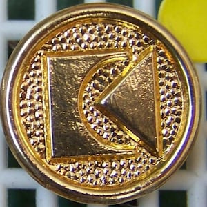 May include: Gold-colored metal button with a geometric design. The button features a square with a triangle inside, creating a stylized letter K.