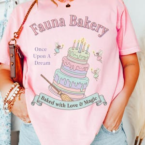 Op de afbeelding: Roze T-shirt met de tekst "Fauna Bakery" en een grillig taartontwerp. De taart heeft meerdere lagen met kaarsen, en het shirt bevat ook de tekst "Once Upon A Dream" en "Baked with Love & Magic".