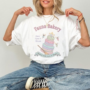 Op de afbeelding: Wit T-shirt met een grillig "Fauna Bakery" ontwerp. De afbeelding toont een meerlagige taart met kaarsen, fee&euml;n en de tekst "Once Upon A Dream" en "Baked with Love & Magic".