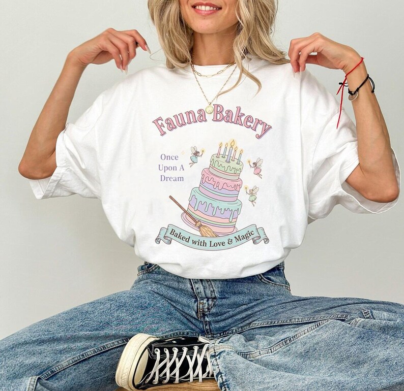 Op de afbeelding: Wit T-shirt met een grillig "Fauna Bakery" ontwerp. De afbeelding toont een meerlagige taart met kaarsen, fee&euml;n en de tekst "Once Upon A Dream" en "Baked with Love & Magic".