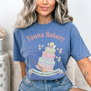 Op de afbeelding: Blauw T-shirt met de tekst "Fauna Bakery" in roze, met een grillig taartontwerp met fee&euml;n en kaarsen. Het shirt heeft ook de tekst "Once Upon A Dream" en "Baked with Love & Magic".