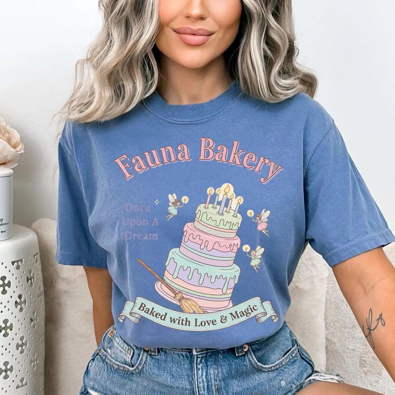 Op de afbeelding: Blauw T-shirt met de tekst "Fauna Bakery" in roze, met een grillig taartontwerp met fee&euml;n en kaarsen. Het shirt heeft ook de tekst "Once Upon A Dream" en "Baked with Love & Magic".
