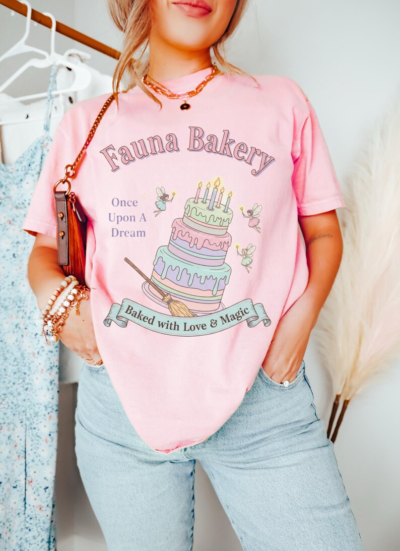 Op de afbeelding: Roze T-shirt met de tekst "Fauna Bakery" en een grillig taartontwerp. De taart heeft meerdere lagen met kaarsen, en het shirt bevat ook de tekst "Once Upon A Dream" en "Baked with Love & Magic".