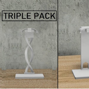 以下が含まれることがあります： 白いヘッドホンスタンドの3個パック。スタンドはプラスチック製で、デザインが異なります。スタンドは木製の表面にあります。