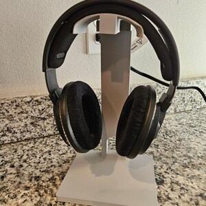 May include: Black over-ear headphones with plush earcups rest on a white headphone stand. The stand has a rectangular base and a vertical support. The headphones are connected to a black cable.