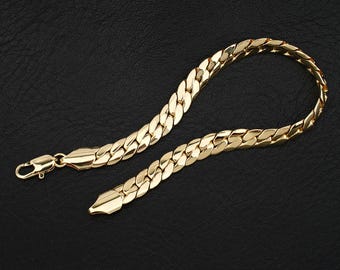 May include: A gold-toned curb chain bracelet featuring interlocking flat, polished links. The bracelet includes a secure lobster claw clasp at one end and a smooth, tapered metal end piece at the other, showcasing a reflective metallic finish.