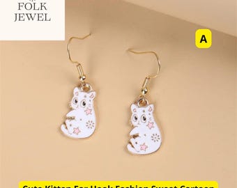 Puede incluir: Un par de pendientes de gato dorados con un acabado de esmalte blanco y detalles en rosa. El gato está sentado con las patas cruzadas y tiene un patrón de estrellas en su cuerpo. Los pendientes están sobre un fondo blanco.