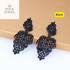 Może przedstawiać: Para czarnych, ozdobnych kolczyków wiszących. Kolczyki mają delikatny, ażurowy wzór z motywami kwiatowymi i zwijanymi. Słowo "Black" jest wyświetlane w żółtym polu. Logo marki "FOLK JEWEL" znajduje się w lewym górnym rogu.