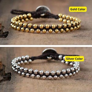 Puede incluir: Dos pulseras con una doble fila de cuentas. La pulsera de arriba tiene cuentas doradas y la pulsera de abajo tiene cuentas plateadas. Ambas pulseras tienen un cordón marrón y un cierre redondo de metal.