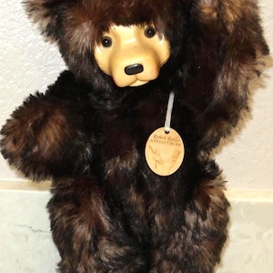 May include: A plush teddy bear with dark brown faux fur, a smooth light-colored wooden face, and black eyes. A round wooden tag hangs from its neck by a ribbon, featuring the text Robert Raikes COLLECTIBLES and an illustration of a hand.