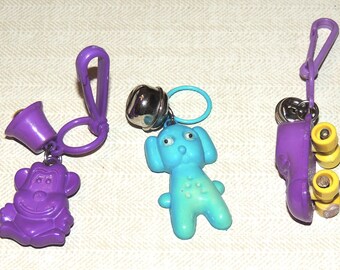 May include: A collection of three novelty keychains. One is a purple monkey with a purple horn, another is a blue dog with a silver bell, and the last is a purple roller skate with yellow wheels.