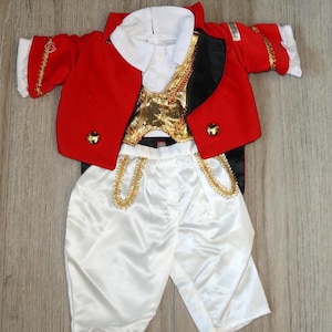 May include: A child's costume featuring a red jacket with gold trim and buttons, a white collared shirt, a gold vest, and white satin pants with gold detailing. The outfit is designed to resemble a military or historical uniform.