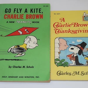 May include: Two vintage Peanuts paperback books. The green book is titled Go Fly a Kite, Charlie Brown by Charles M. Schulz, featuring a red kite illustration. The yellow book is titled A Charlie Brown Thanksgiving by Charles M. Schulz.