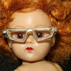May include: A red-haired doll wearing silver-rimmed glasses with a geometric design. The doll has a porcelain face with painted red lips and closed eyes.