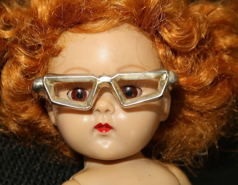 May include: A red-haired doll wearing silver-rimmed glasses with a geometric design. The doll has a porcelain face with painted red lips and closed eyes.