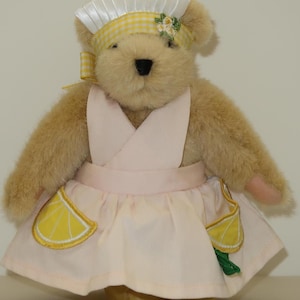May include: A plush teddy bear wearing a pink dress with yellow lemon accents and a white and yellow gingham headband. The bear is wearing a white hat with a ruffled edge.