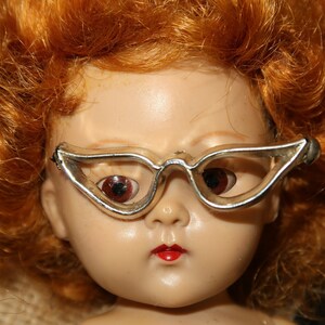 May include: A vintage doll with red hair wearing silver cat-eye glasses. The doll has a porcelain face with painted features and red lips.