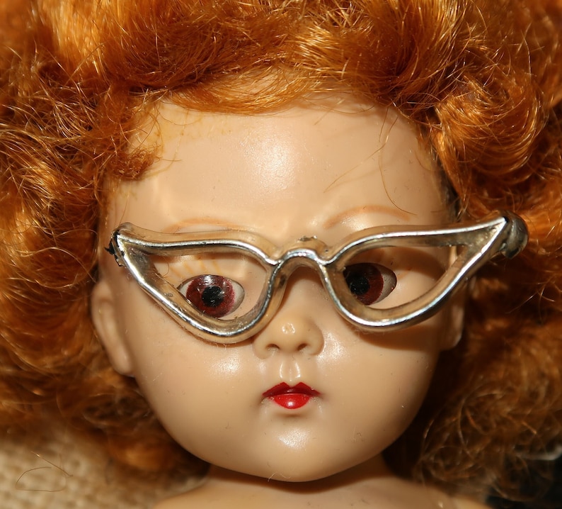 May include: A vintage doll with red hair wearing silver cat-eye glasses. The doll has a porcelain face with painted features and red lips.